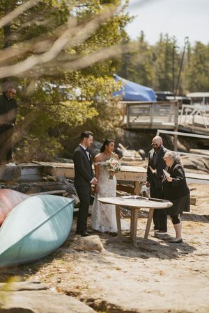 Bride and groom signing wedding documents on a sandy lakeside beach with canoes and a wooden dock in the background.