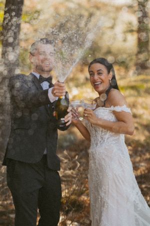 Joyful bride and groom popping champagne bottle outdoors in forest setting on their wedding day.