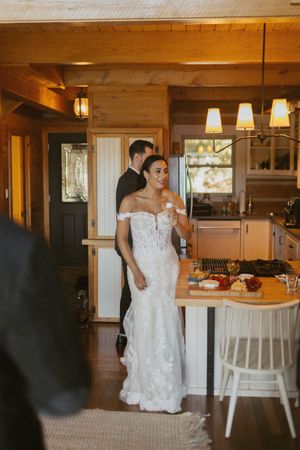 Smiling bride in off-shoulder lace wedding gown holding champagne in a warm wooden cabin kitchen with groom.