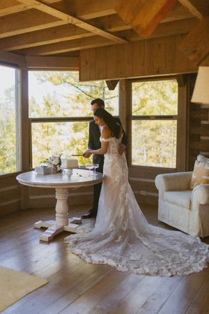 Bride and groom cutting wedding cake at a white pedestal table in a sunlit rustic log cabin interior.