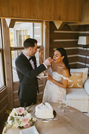 Newlyweds feeding each other wedding cake inside a cozy rustic log cabin with wooden walls and warm lighting.