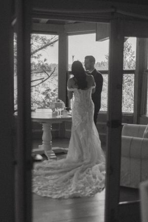 Black and white photo of bride and groom sharing a romantic moment by large windows in a rustic cabin interior.