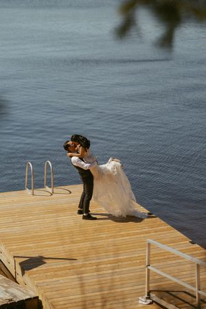 Groom lifting bride for a romantic kiss on a wooden dock overlooking a serene blue lake on their wedding day.