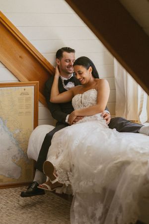 Bride and groom sit together laughing near a staircase beside a framed nautical map in a cozy cottage interior.