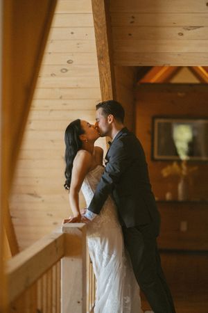 Bride and groom sharing a tender kiss on a wooden balcony inside a warm rustic log cabin wedding venue.