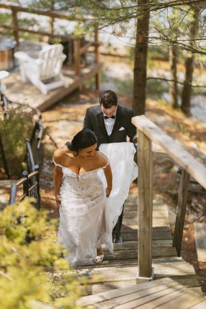 Groom in black tuxedo carries bride's white lace wedding gown train as couple walks up wooden stairs outdoors.