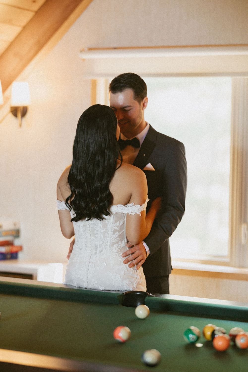 Bride and groom share a romantic kiss near a pool table, dressed in wedding attire in a warmly lit room.