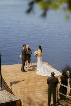 Bride and groom exchange vows on a sunlit wooden dock overlooking a calm blue lake during an intimate outdoor wedding ceremony.