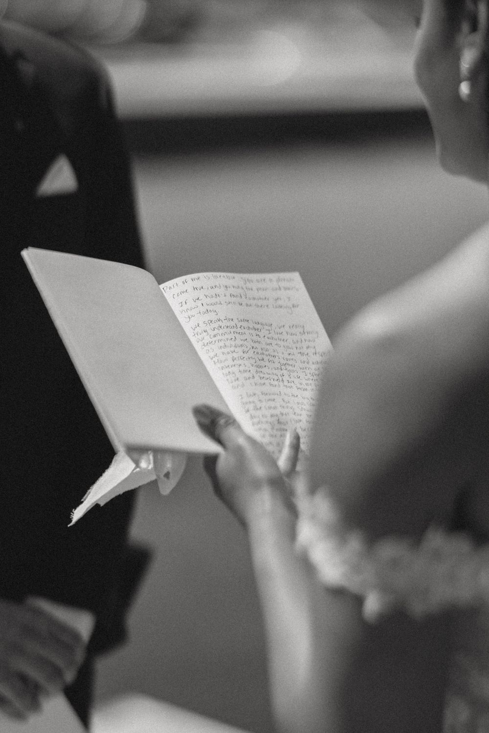 Bride reading handwritten wedding vows from a small booklet during ceremony, black and white photo.