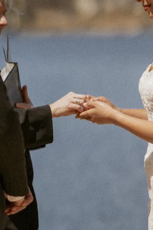 Bride placing a wedding ring on groom's finger during outdoor ceremony near water.