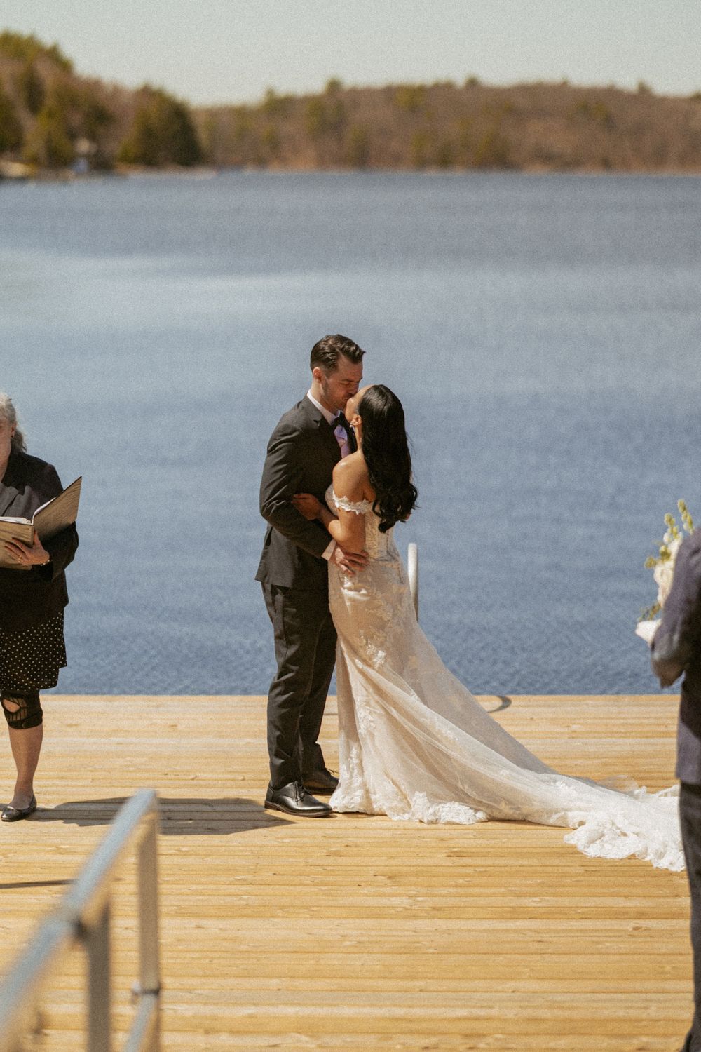 Bride and groom share first kiss on a wooden dock overlooking a serene blue lake during outdoor wedding ceremony.