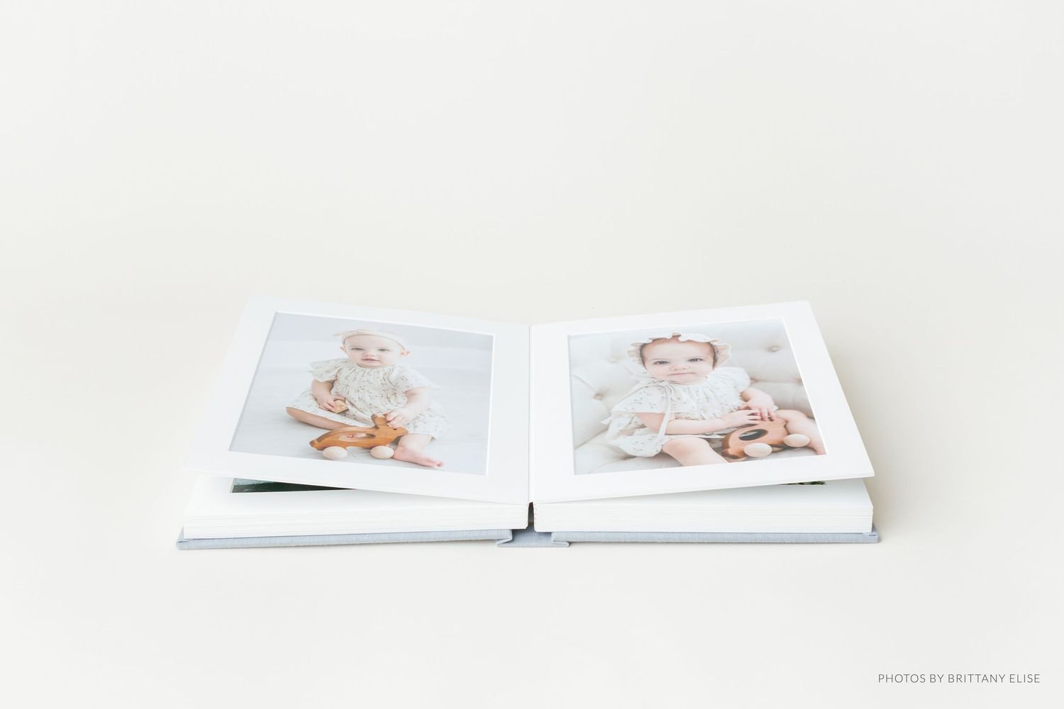 Open photo album displaying two portrait images of a baby girl in white outfits on a light background.
