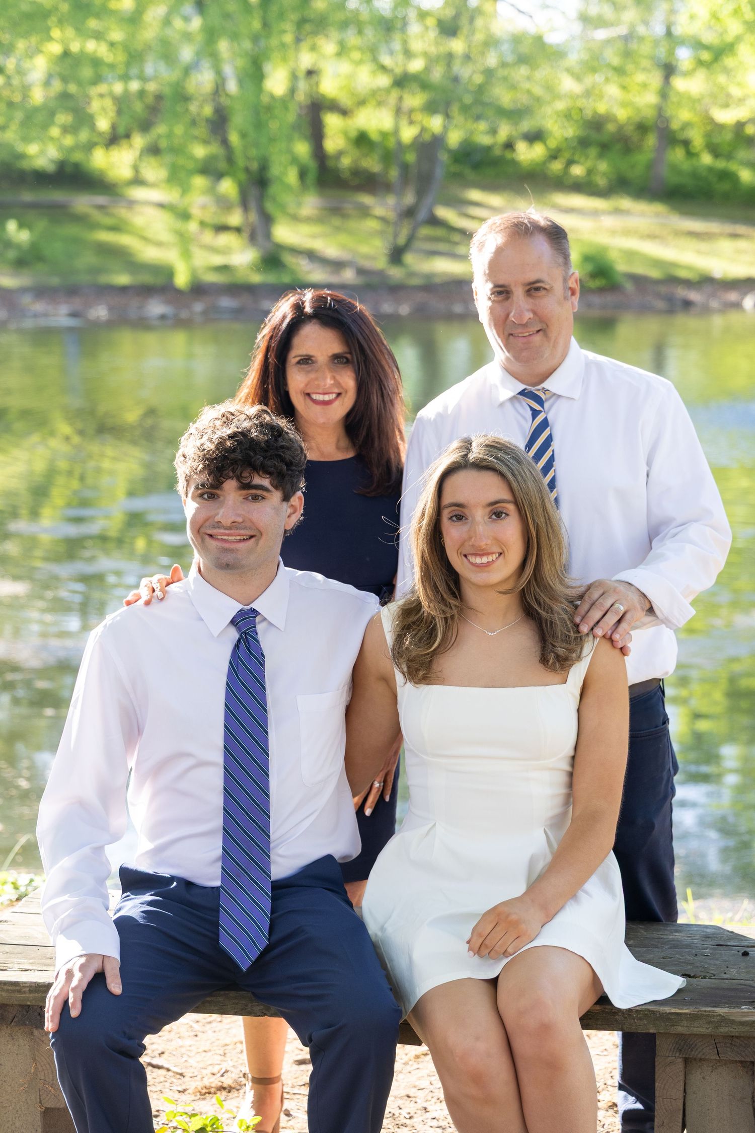 Family of four posing outdoors near a pond, two seated in front, two standing behind, dressed in formal attire.