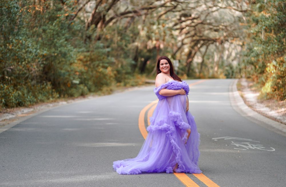 Pregnant woman in flowing lavender gown poses on tree-lined road for elegant maternity photography session.