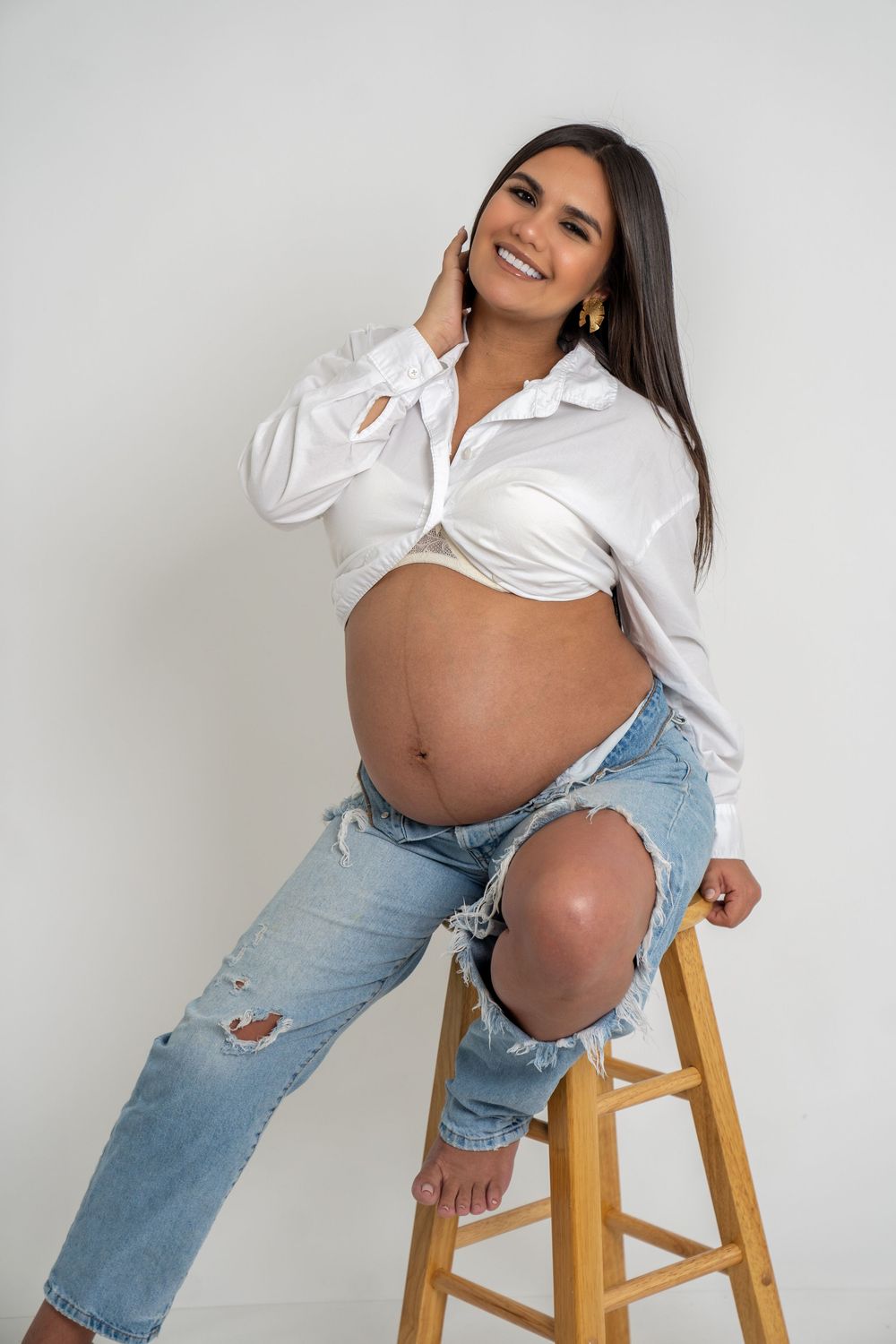 Smiling pregnant woman in white shirt and ripped jeans sitting on wooden stool against white background.