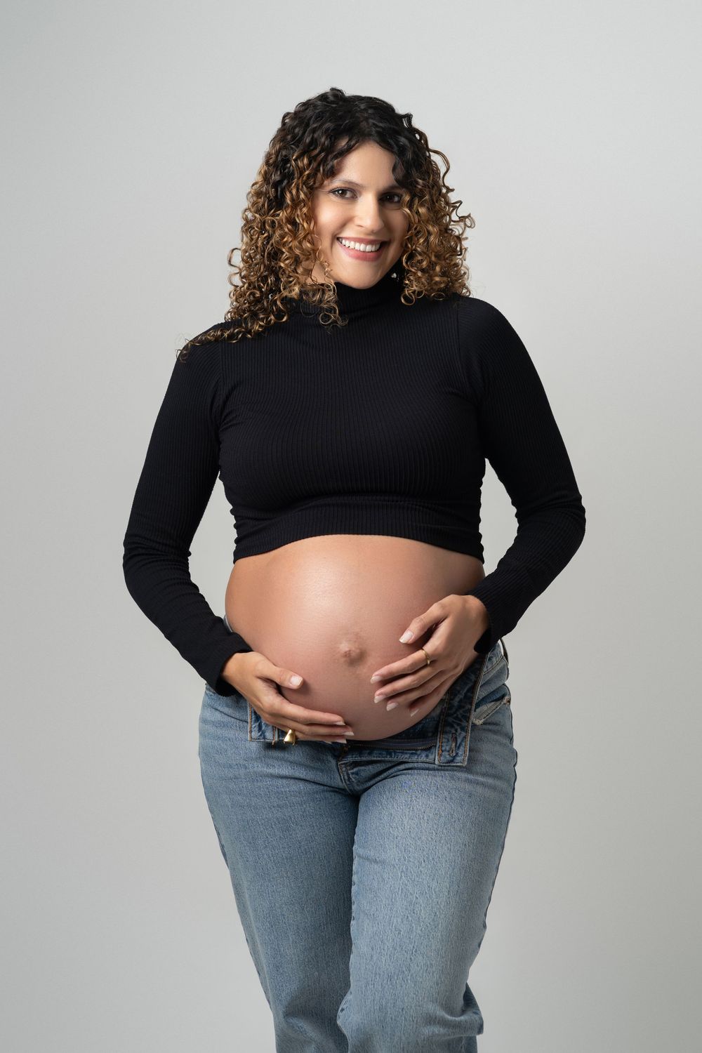 Smiling pregnant woman with curly hair wearing black crop top and jeans, cradling her bare baby bump.