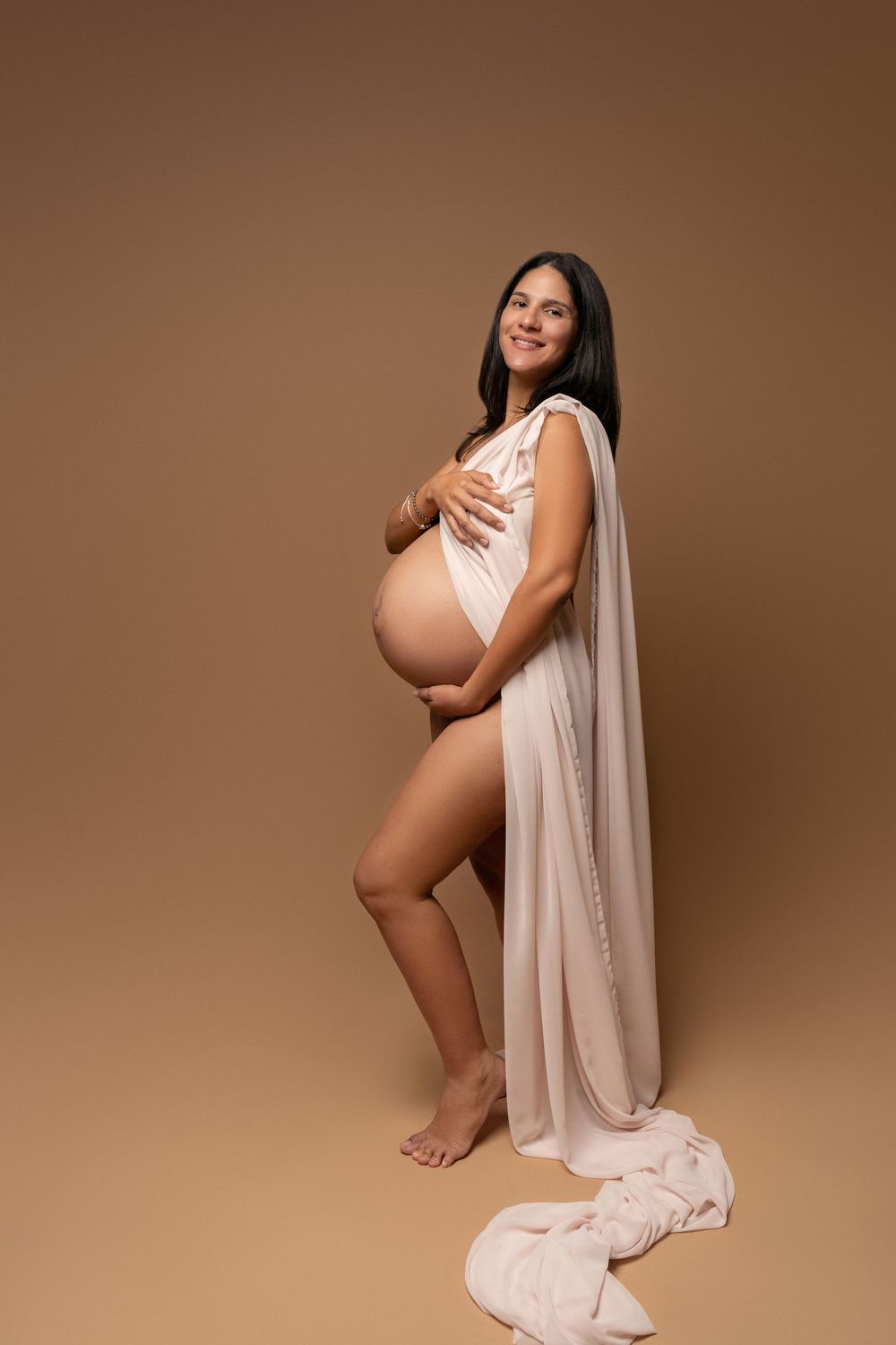 Smiling pregnant woman in sheer beige fabric wrap cradling her baby bump, posing barefoot on a warm brown background.