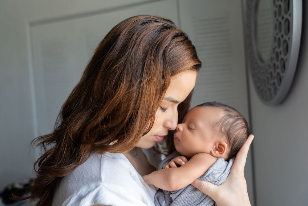 A mother tenderly touches foreheads with her sleeping newborn baby wrapped in a gray swaddle.