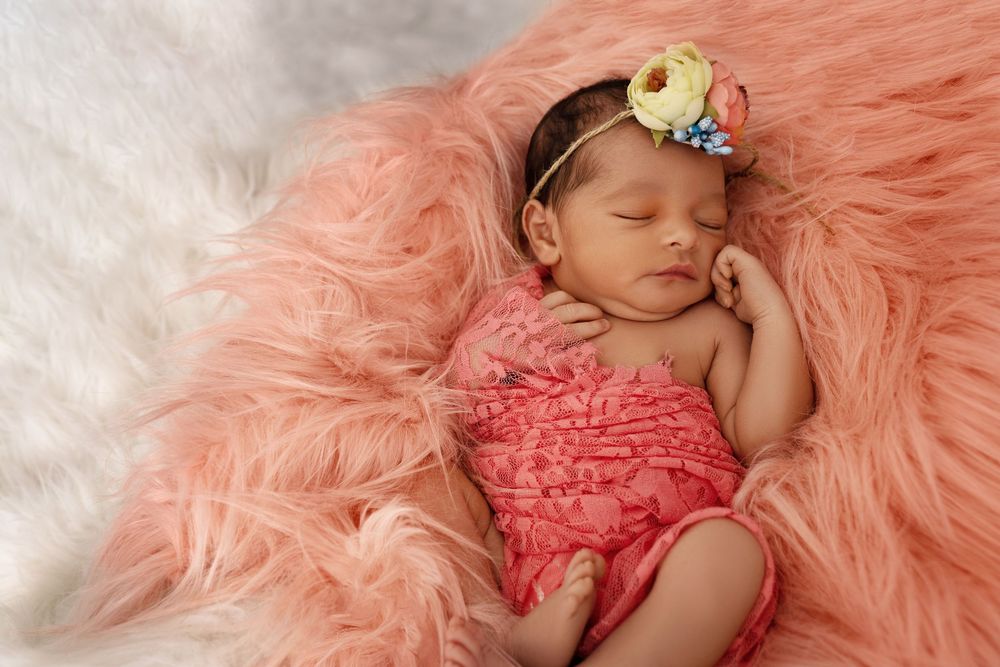 Sleeping newborn baby girl in pink wrap and floral headband on fluffy peach fur backdrop.