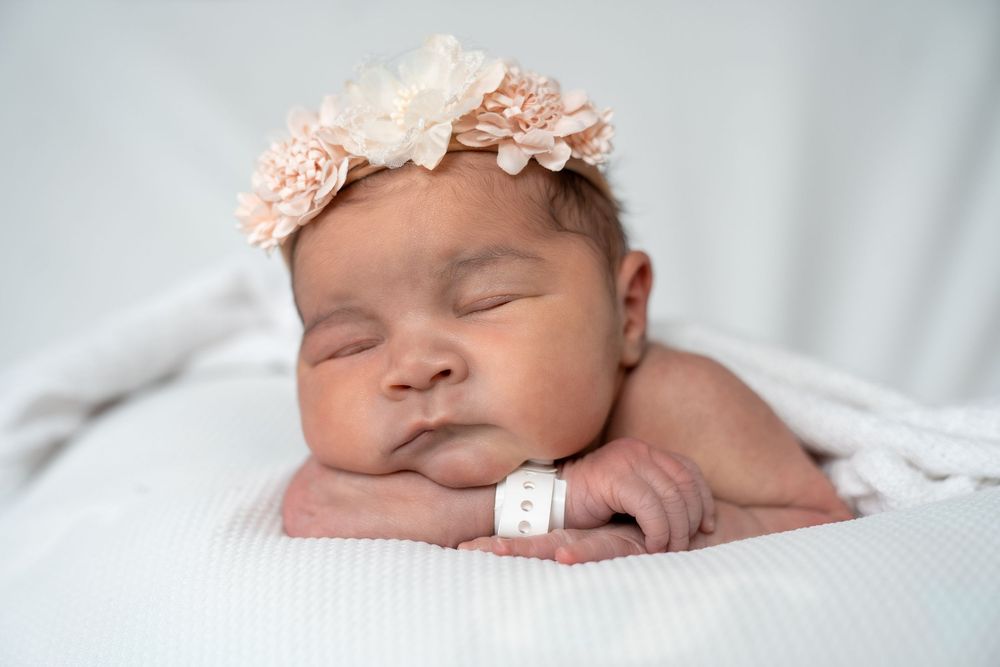 Sleeping newborn baby girl wearing a pink floral headband, resting on a white blanket with a hospital wristband.