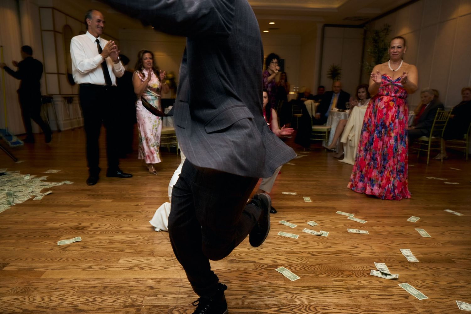 Man dancing energetically on a wooden dance floor at a wedding reception with guests watching and money scattered around.