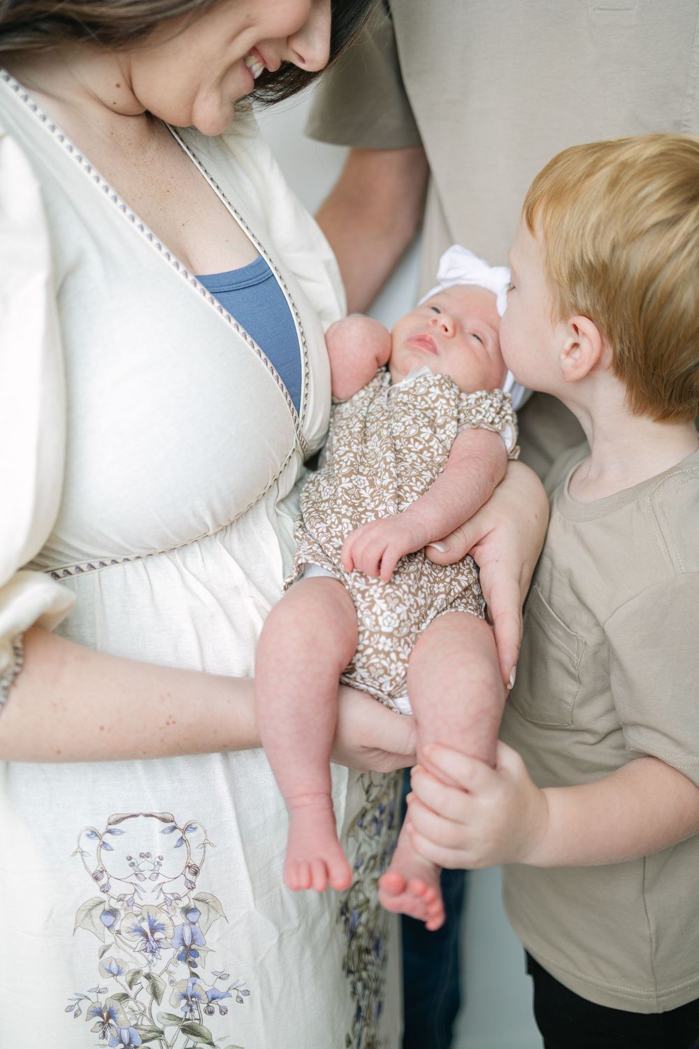 A mother in a white dress holds her newborn baby girl while her young son gives the infant a gentle kiss.