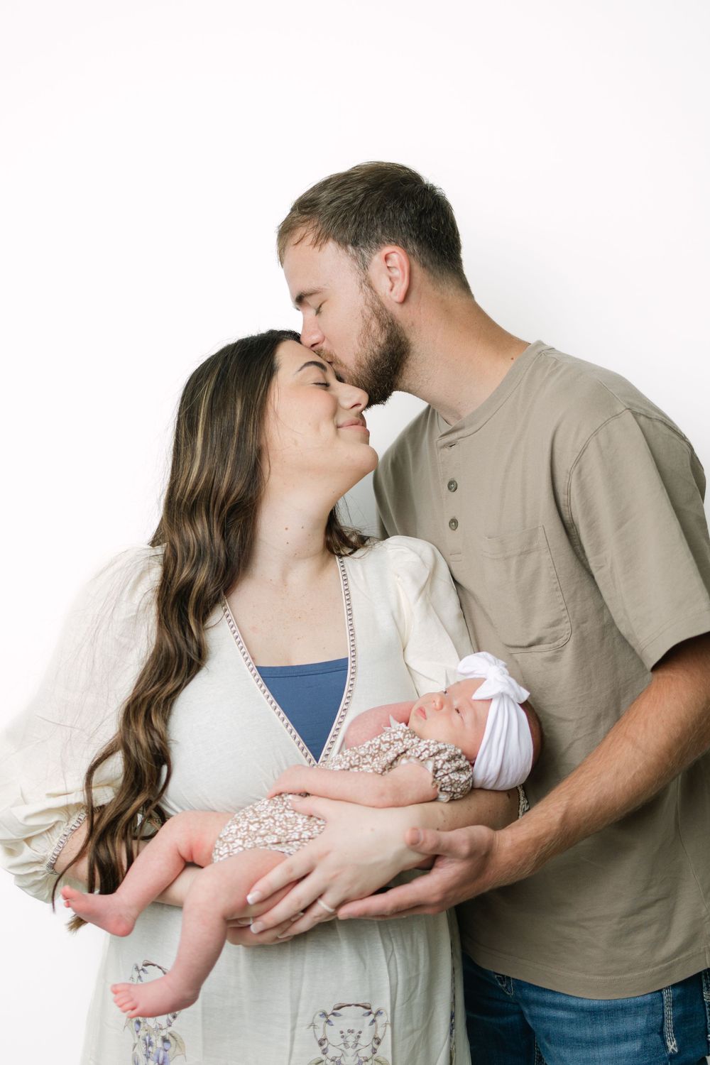 New parents share a tender kiss while cradling their swaddled newborn baby together against a white background.