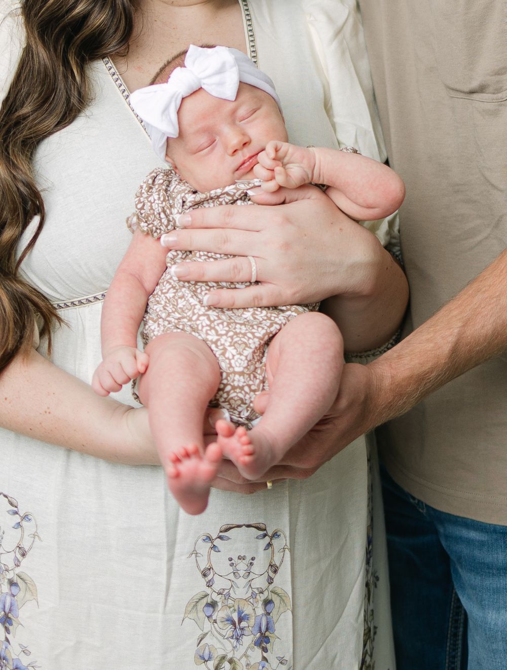 Parents cradling their sleeping newborn baby girl in a white bow headband and floral romper during a newborn photo session.