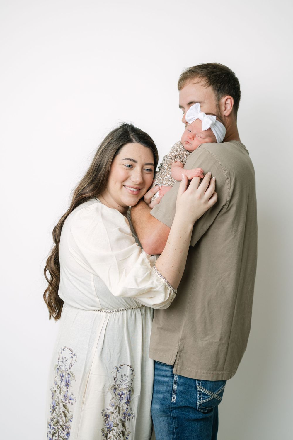 Happy couple in neutral tones holding their newborn baby girl with a white bow against a white background.