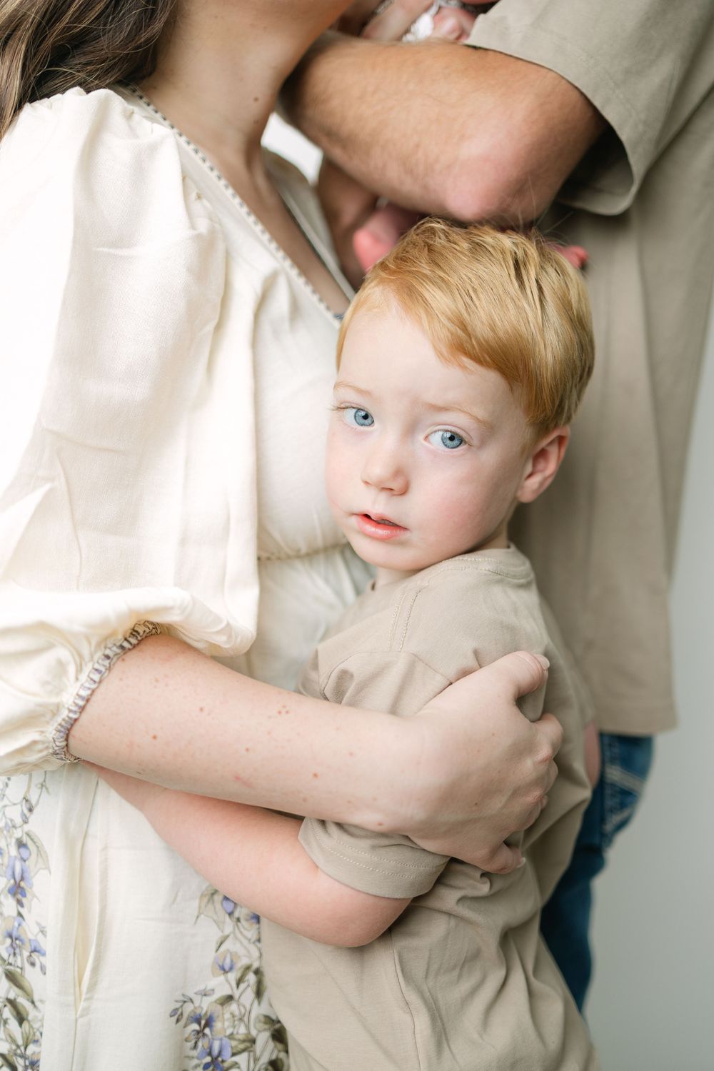 A red-haired blue-eyed toddler being held by his mother in a white floral dress during a family portrait session.