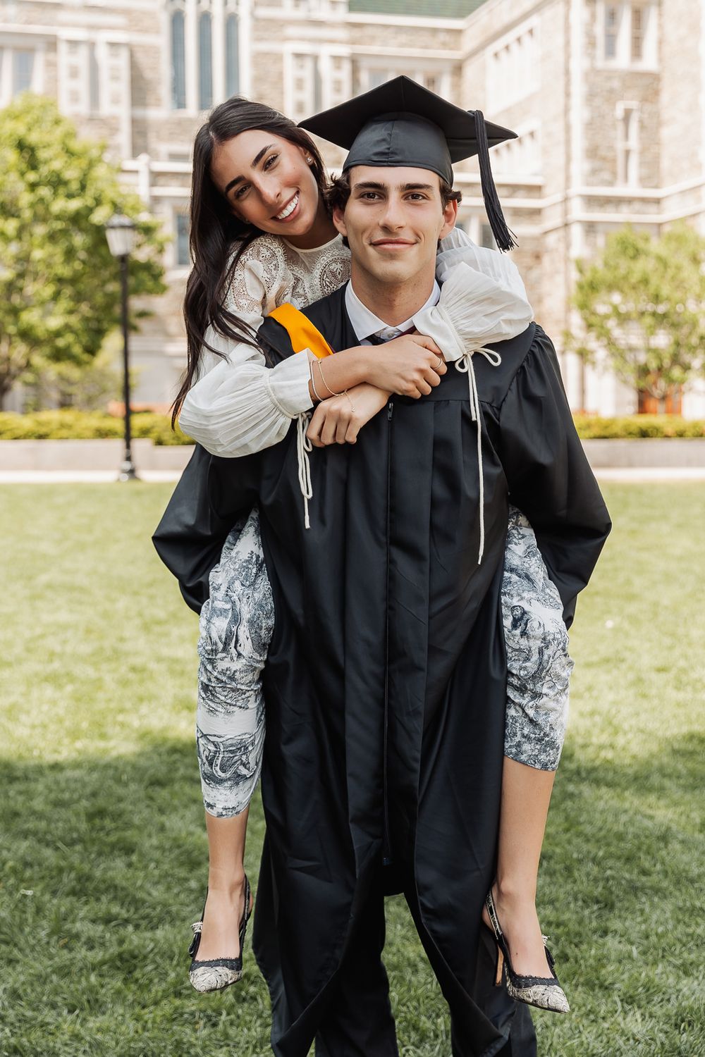 A young man in graduation cap and gown gives a smiling woman a piggyback ride on a green campus lawn.