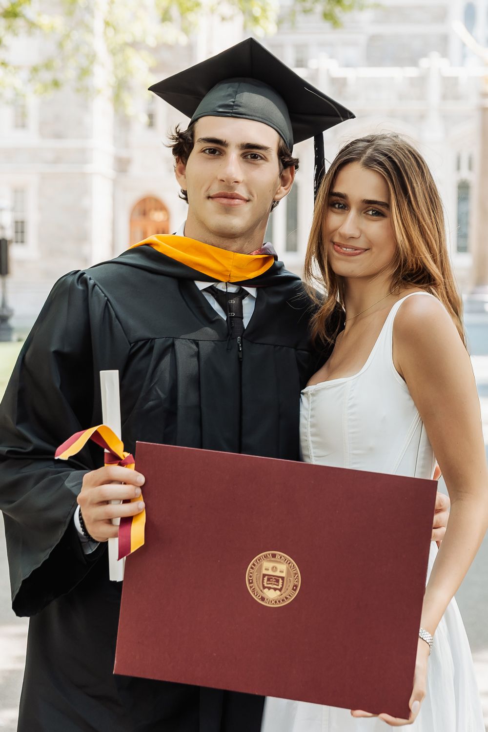 Young male graduate in cap and gown holds diploma with female companion in white dress outdoors.