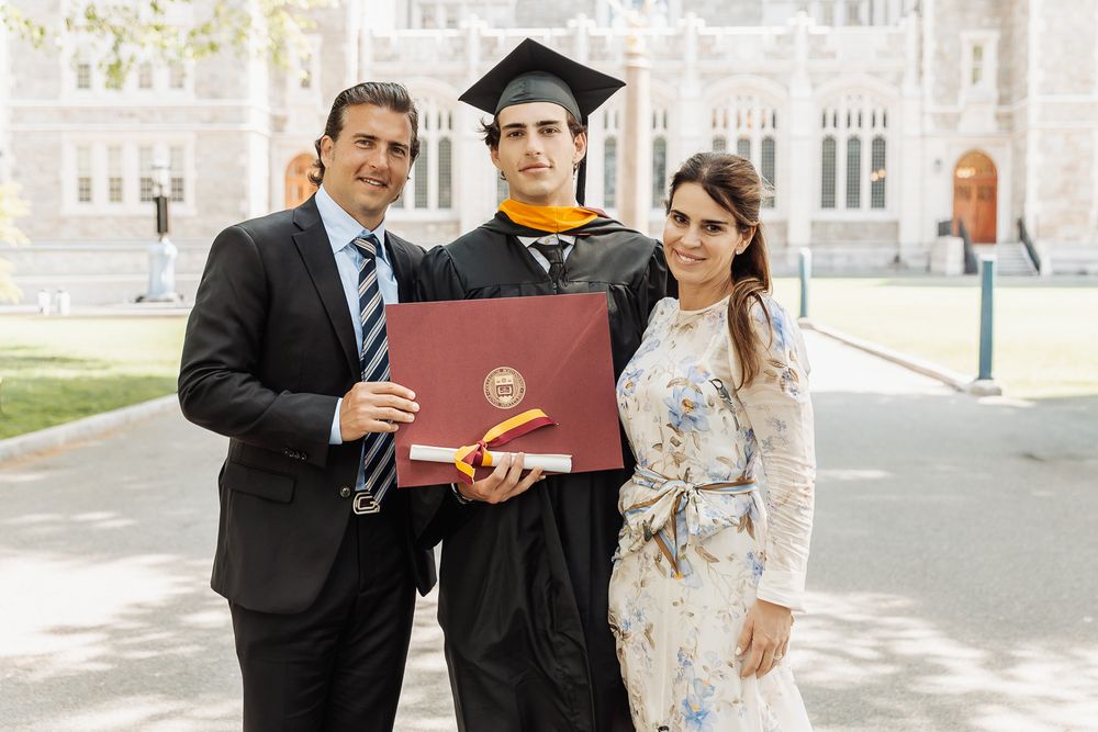 Graduate in cap and gown holds diploma with proud parents at outdoor university graduation ceremony.