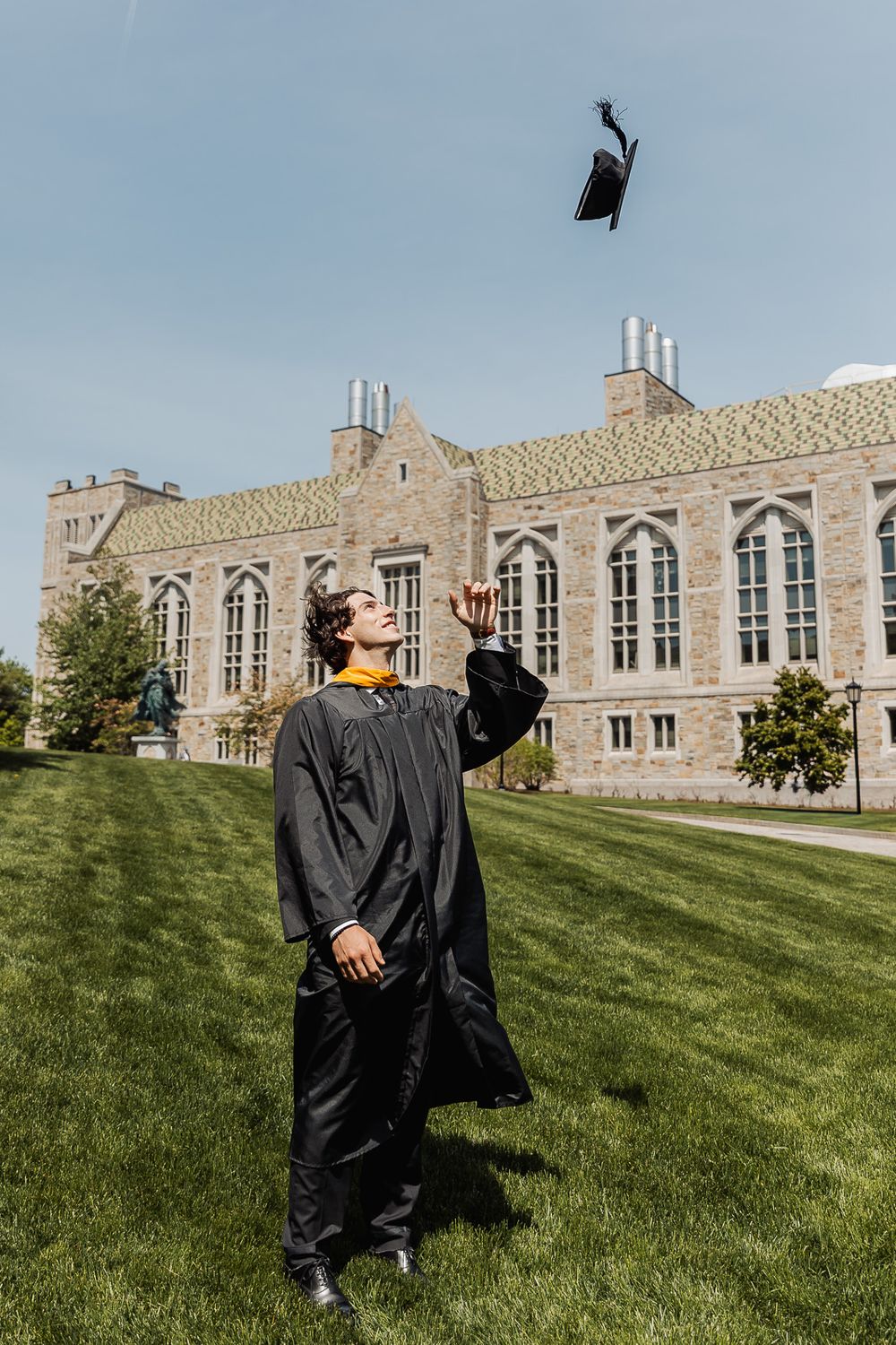 Graduate in black gown tosses cap in air outside historic stone university building on sunny day.