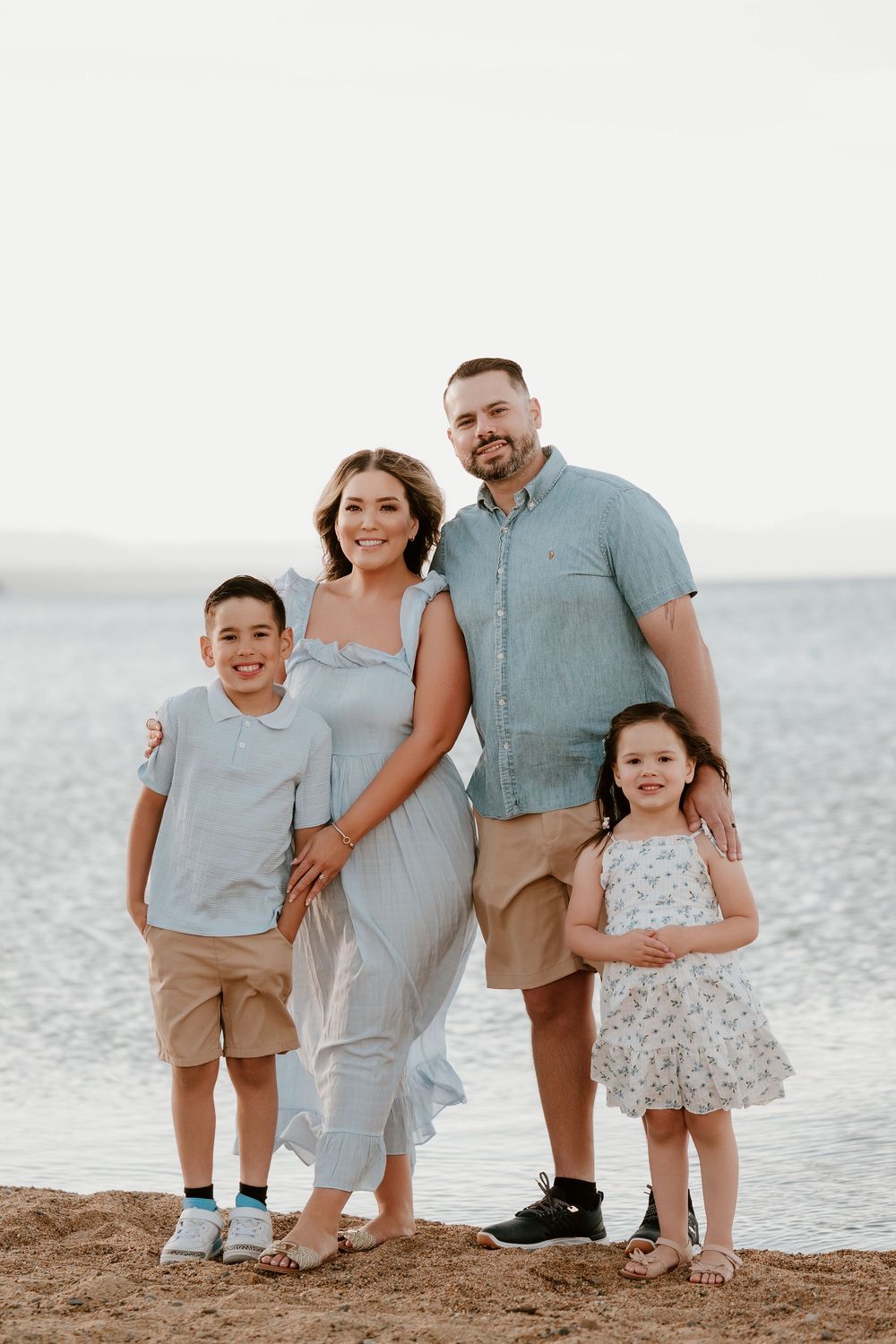 Family of four smiling together on a sandy beach, parents with young boy and girl in coordinating blue and khaki outfits.