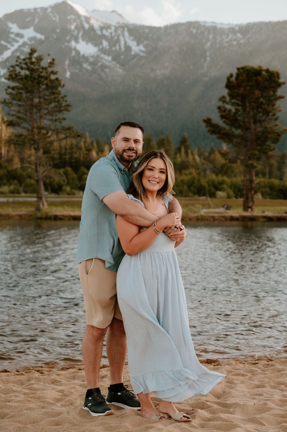 Couple embracing on sandy beach with snow-capped mountains and lake backdrop.