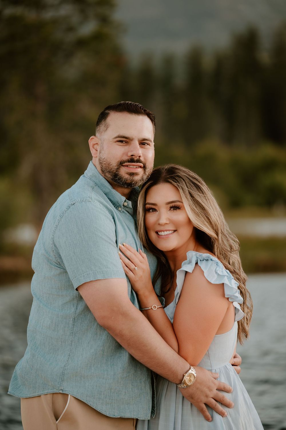 A smiling couple embraces outdoors near a lake with pine trees, wearing coordinating blue outfits in a romantic portrait.