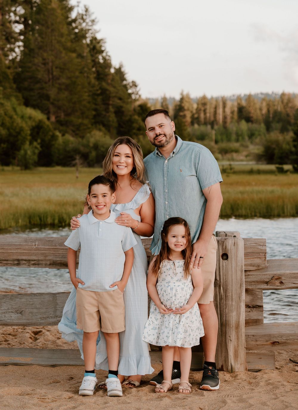 Happy family of four posing outdoors near a lake and green trees on a sunny day.