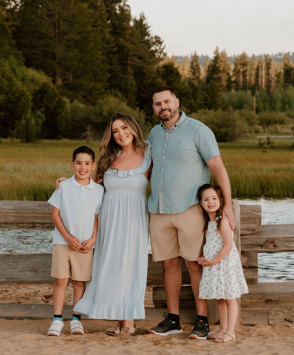 A happy family of four poses outdoors near a lake, dressed in light summer clothing, surrounded by green trees.