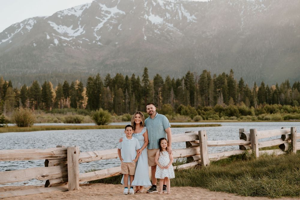 Family of four posing by a wooden fence near a mountain lake surrounded by pine trees and snow-capped peaks.
