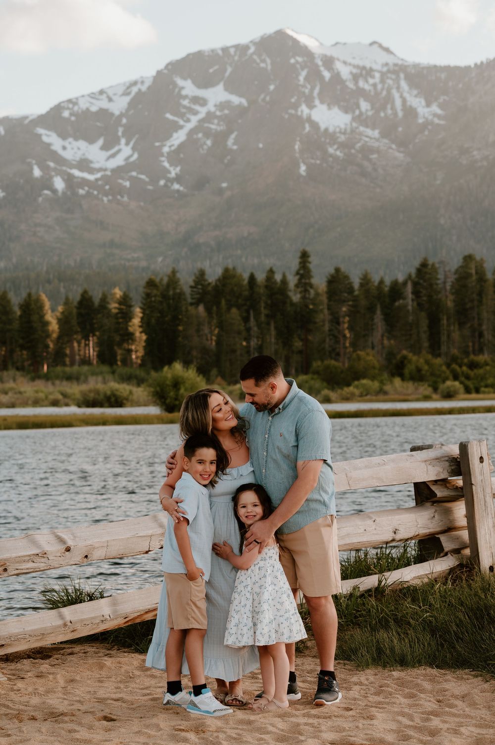 Family of four posing on beach with snow-capped mountains and lake behind them.