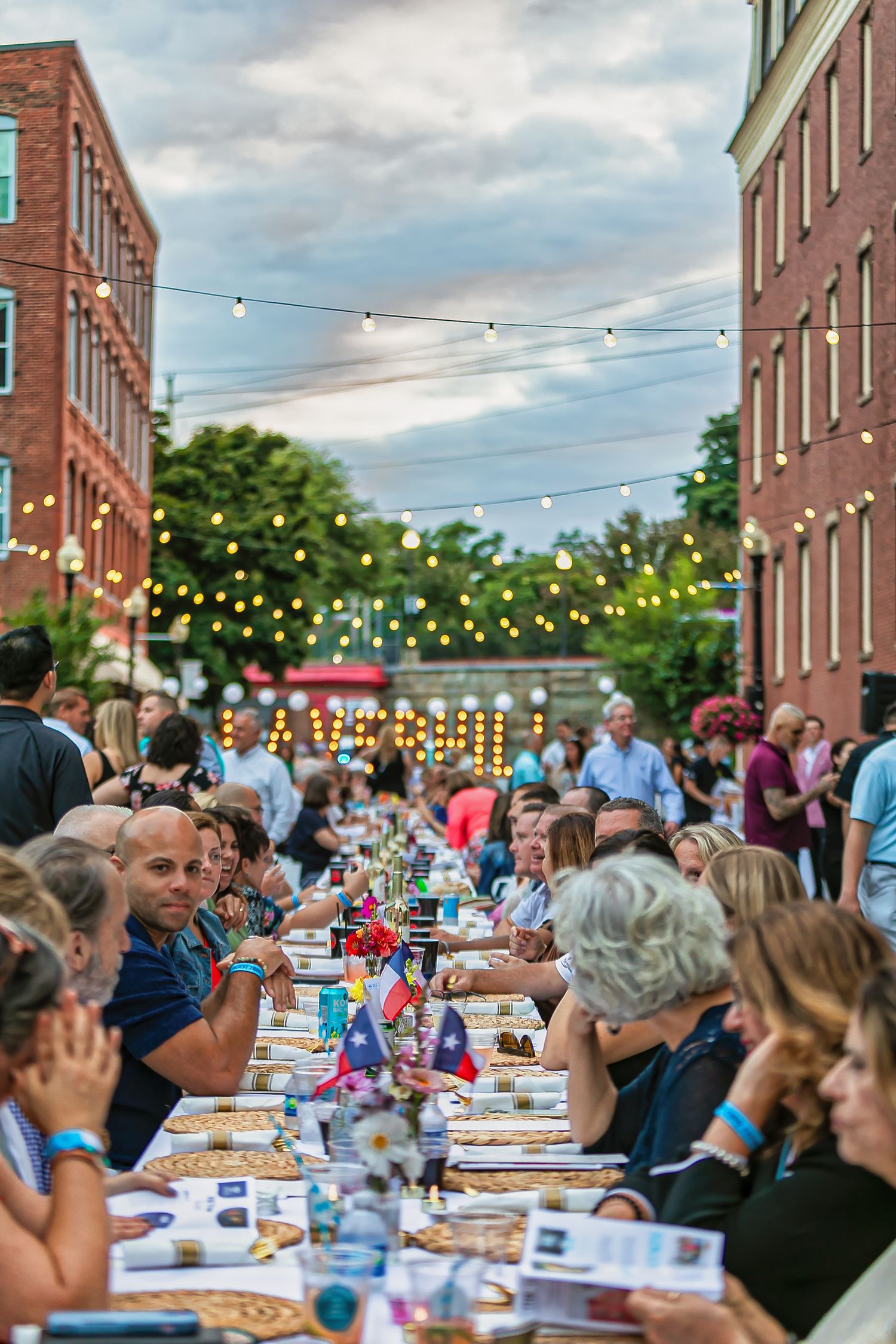 Outdoor street dining event with long tables, string lights, and crowds of people enjoying food between brick buildings at dusk.