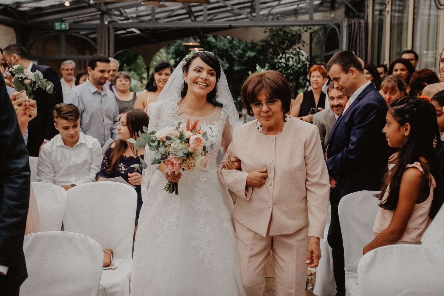 Bride in white gown walks down aisle with older woman in pink suit during indoor wedding ceremony.