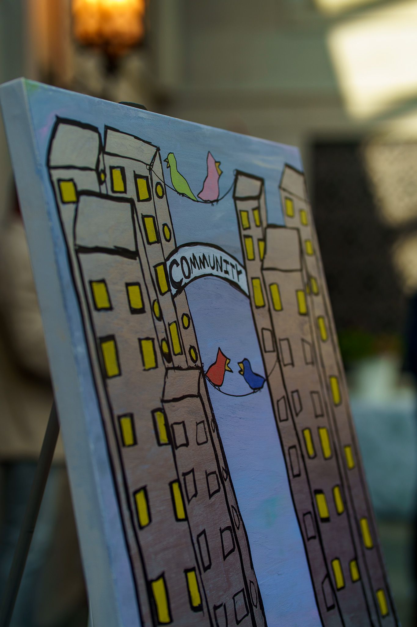 Painting on easel depicting two towers with arch reading Community, colorful birds flying above, yellow windows.