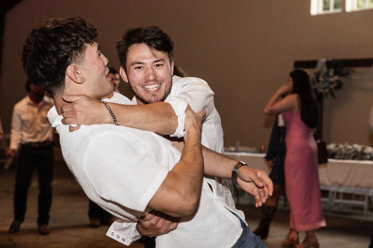 Two men dancing and laughing together at a wedding reception celebration indoors.