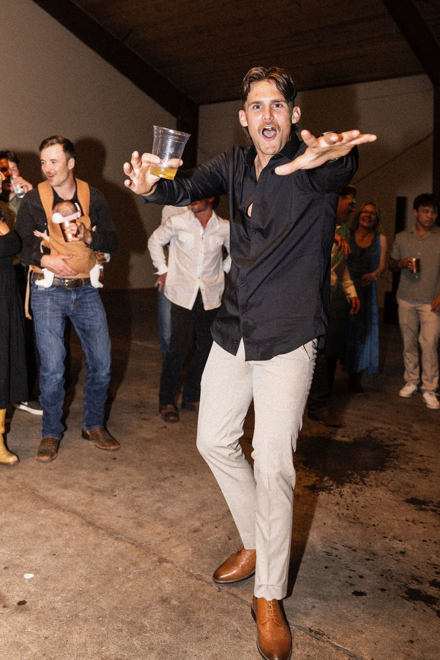 Energetic man in black shirt and khakis dancing at a party, holding a plastic cup of beer.