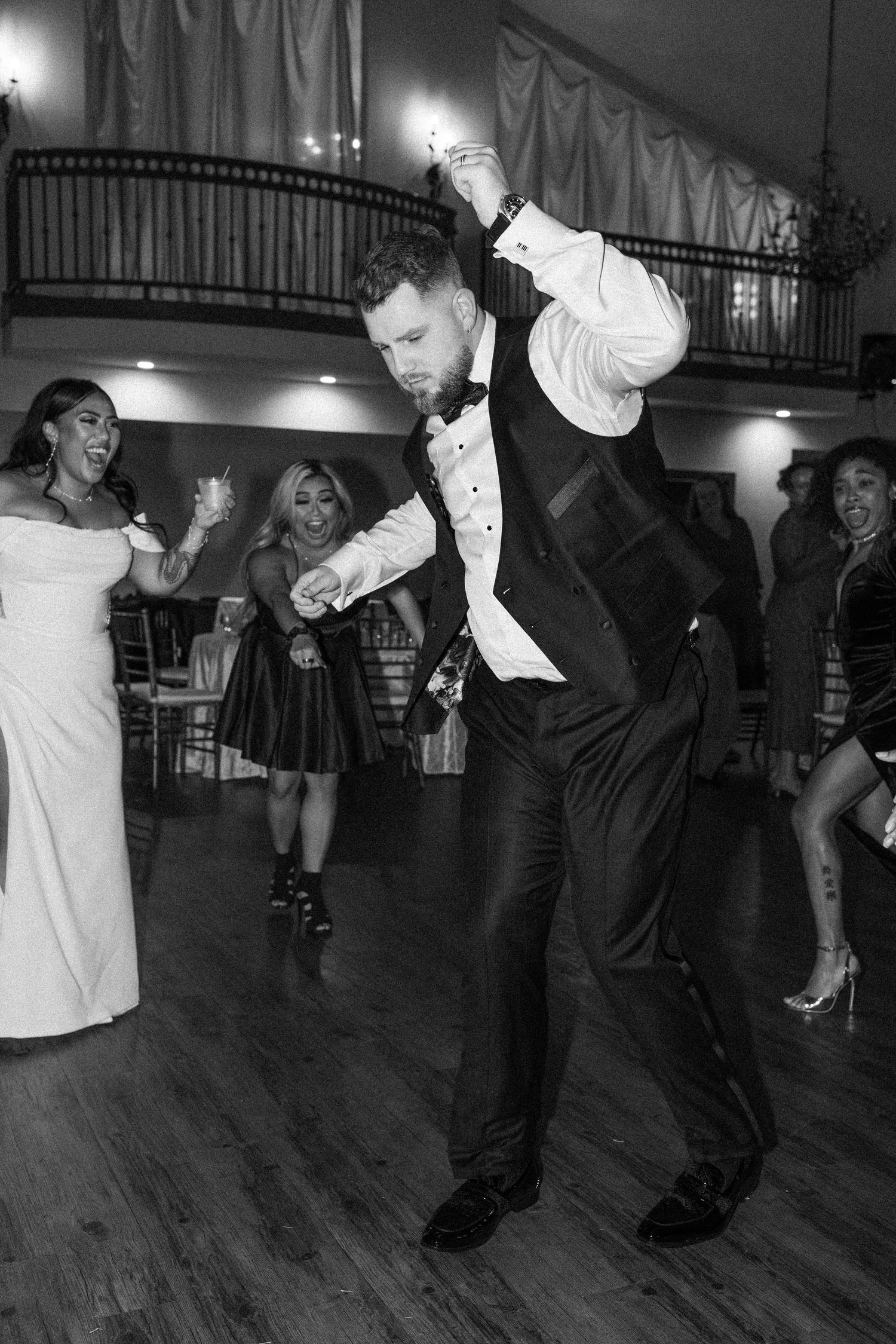 A groom dances energetically at his wedding reception while guests celebrate in the background in this black and white photo.