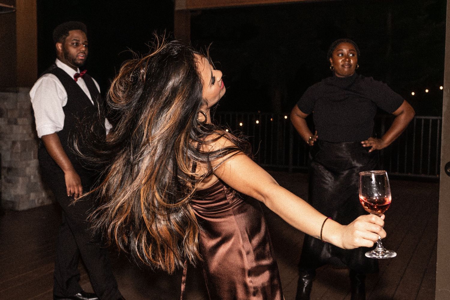 Woman in brown dress dancing with wine glass at elegant evening event with guests in background.