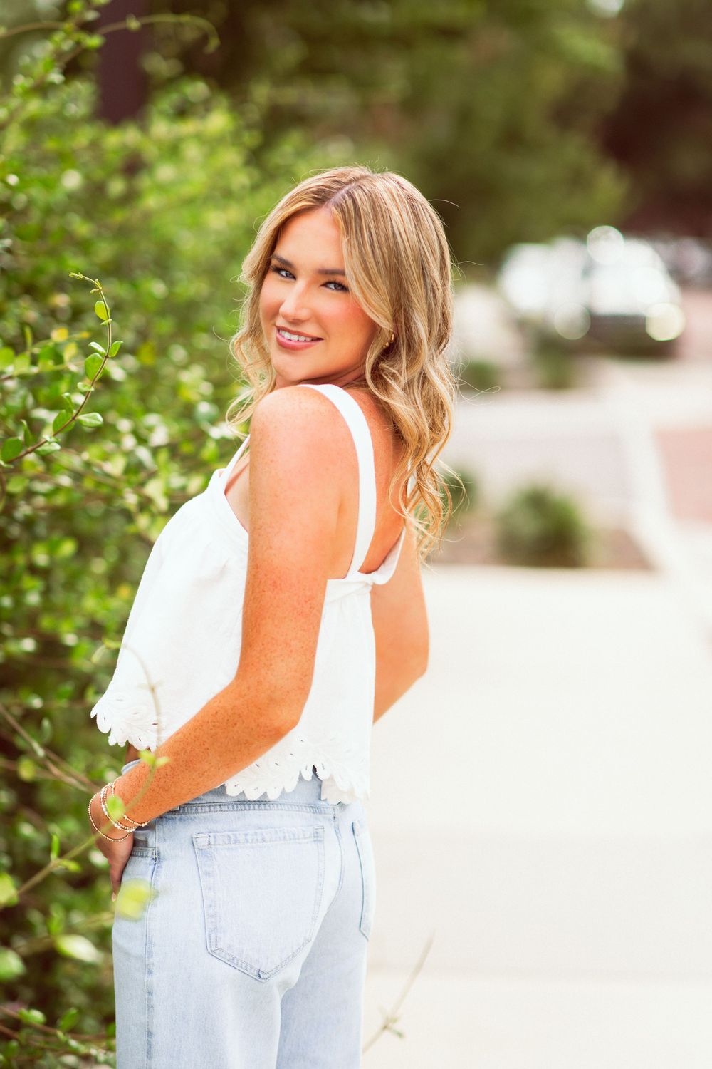 Blonde woman in white crop top and light jeans smiling over shoulder outdoors.