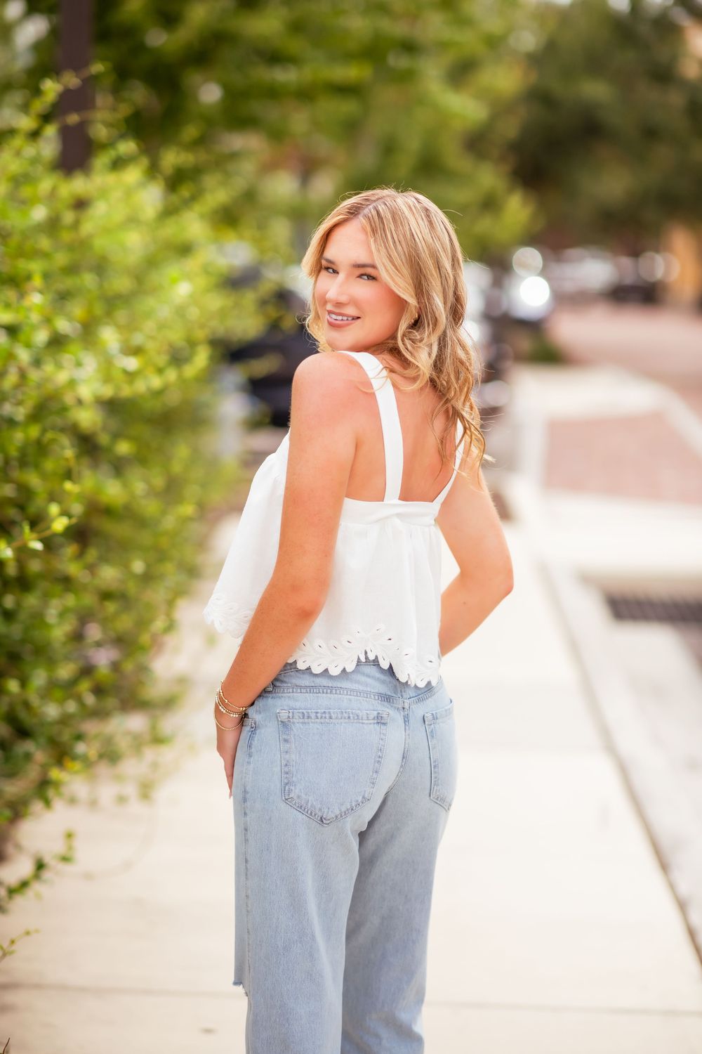 Blonde woman in white lace crop top and jeans posing on a sunny sidewalk.